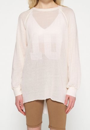 Kobieta ubrana w luźny, długi sweter z białej siateczki na białej koszulce na ramiączkach i dopasowane brązowe szorty kolarskie.