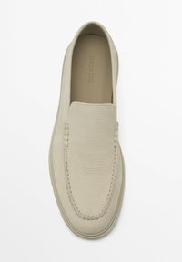 Mocassin beige en cuir à enfiler avec un dessus texturé et une couture visible le long de la pointe arrondie sur un fond blanc.