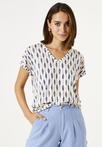 Leichtes, kurzärmeliges Oberteil mit V-Ausschnitt, das eine beige Basis und ein blaues Blattmuster aufweist. Kombiniert mit hellblauen, taillierten Hosen.