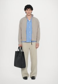 Beige lightweight jacket over a blue button-up shirt, paired with light trousers and a black leather tote bag, set against a plain background.