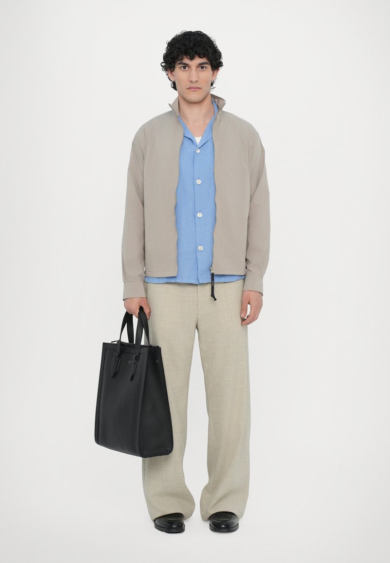 Beige lightweight jacket over a blue button-up shirt, paired with light trousers and a black leather tote bag, set against a plain background.