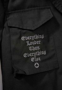 Černá látková kapsa košile se šedým vyšitým textem: „Everything Louder Than Everything Else“ s ikonou pikové káry pod ním.