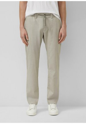 Person, die beigen, gerade geschnittenen Hosen mit Kordelzug an der Taille, weiße Turnschuhe und ein in die Hose gestecktes weißes T-Shirt vor einem schlichten Hintergrund trägt.