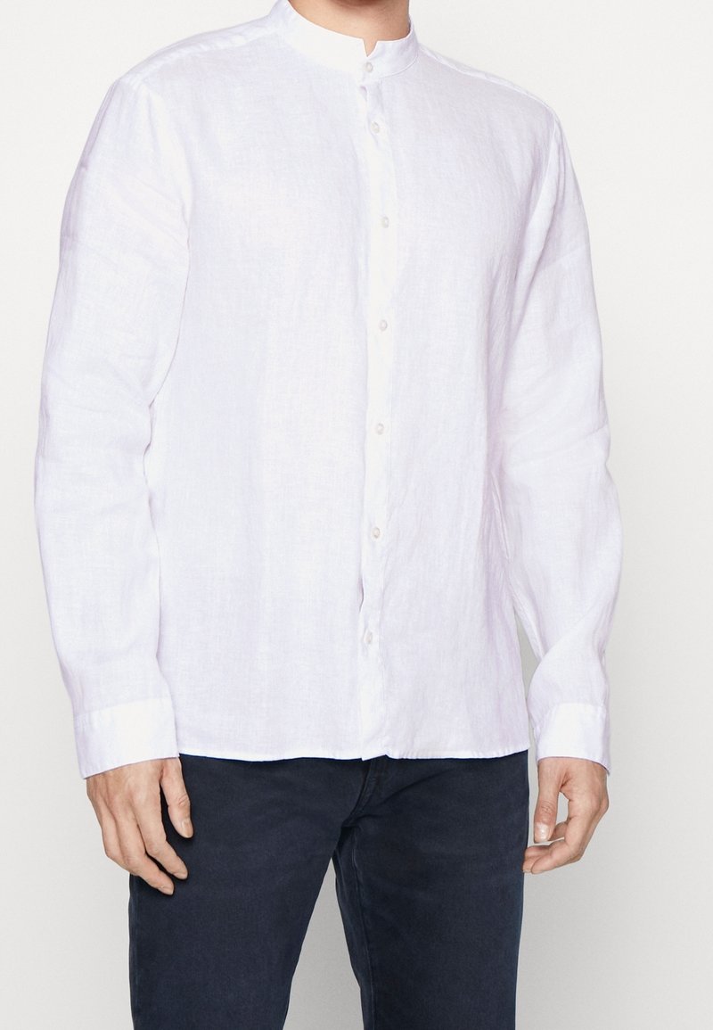 Homme portant une chemise blanche à manches longues avec col mandarin et boutons, ainsi qu’un pantalon noir, visible du cou jusqu’aux hanches.