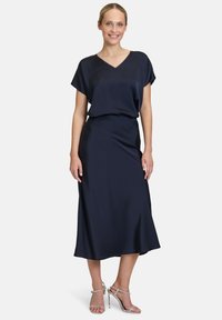 Abito in raso blu navy con scollatura a V, maniche corte e una silhouette fluida che arriva a metà polpaccio. Abbinato a sandali con tacco a strappo argento.