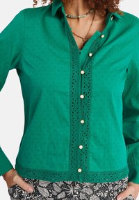 Chemise en coton verte avec boutons, ornée de détails en œillets sur le devant et à l'ourlet, avec des boutons décoratifs et des manches longues.