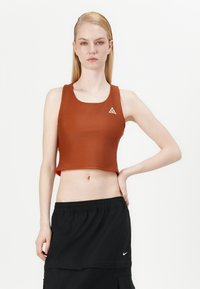 Top corto marrón de material suave, con un pequeño logo blanco en forma de triángulo en el pecho izquierdo, combinado con una falda negra.