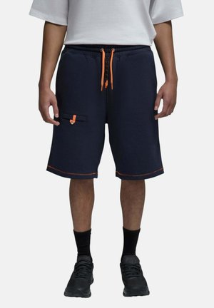 Personne portant un short bleu marine foncé avec des cordons et coutures orange, une chemise blanche, des chaussettes noires et des chaussures de sport noires, debout.
