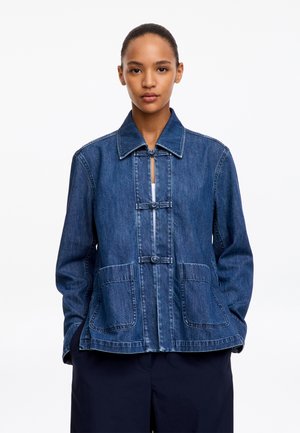 Jeune personne portant une veste en jean bleue avec poches plaquées à l'avant et boutons à bascule par-dessus un haut blanc sur fond uni.