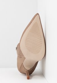 Zapato de tacón alto en color beige, con punta afilada, suela de goma y cremallera en el lateral, ubicado de pie contra una pared blanca en la esquina.