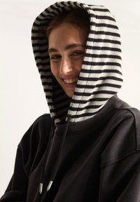 Zwarte sweatshirt met een geribbelde, gestreepte capuchon in zwart en wit. Voorzien van trekkoorden met zilveren metalen uiteinden. Getextureerde stof, relaxte pasvorm.