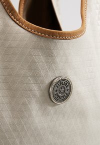 Beige tekstureret håndtaske med brune syninger og et rundt metal Kipling-logo badge på fronten.