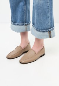 Pantofi slip-on din piele bej cu vârf ascuțit, asociați cu pantalonii din denim albastru, cu tivuri răsfrânte și căptușeală de un albastru deschis, având detalii cu fermoar.