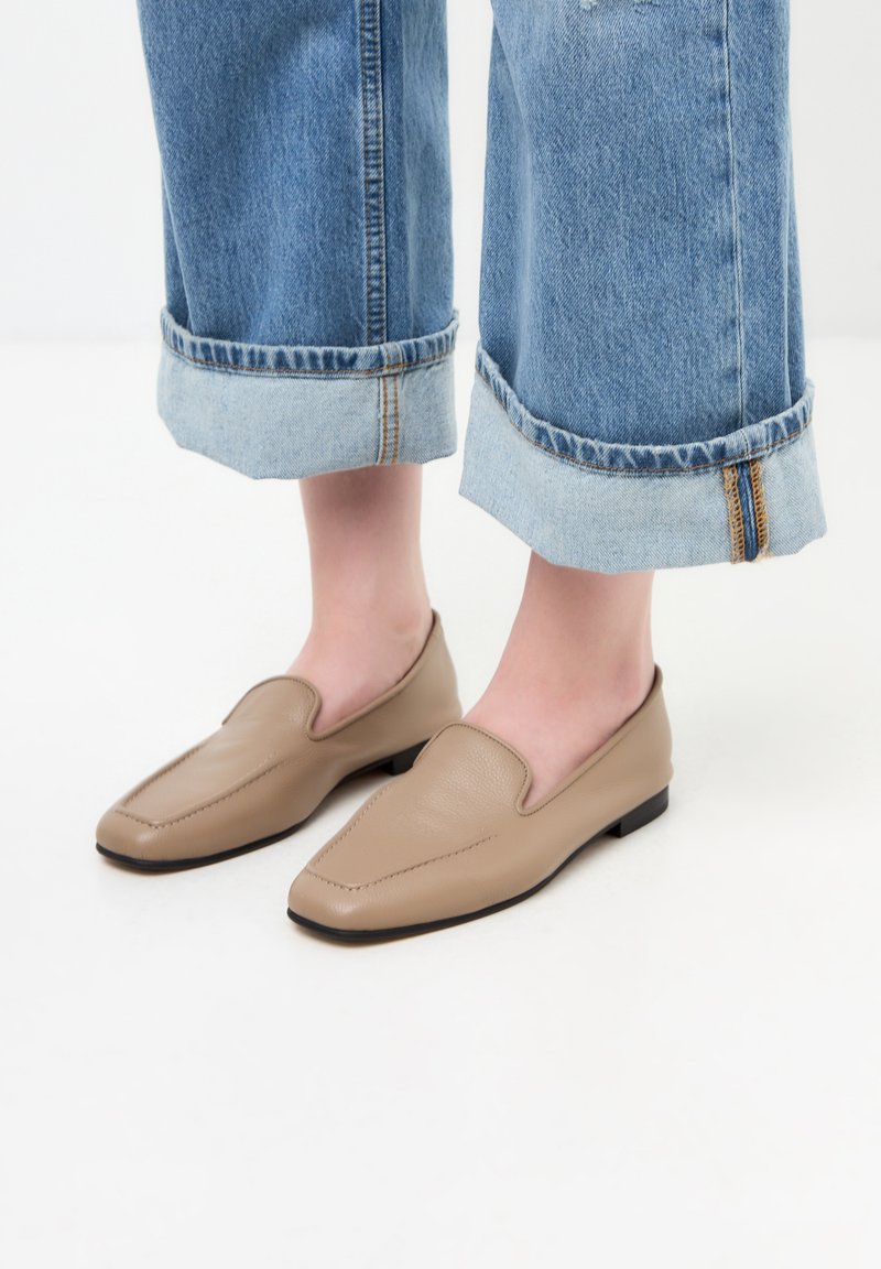 Pantofi slip-on din piele bej cu vârf ascuțit, asociați cu pantalonii din denim albastru, cu tivuri răsfrânte și căptușeală de un albastru deschis, având detalii cu fermoar.