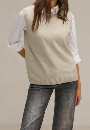 Femme portant un pull beige sans manches par-dessus une chemise blanche à manches longues avec les manches retroussées et un jean noir délavé.