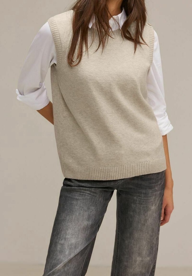 Femme portant un pull beige sans manches par-dessus une chemise blanche à manches longues avec les manches retroussées et un jean noir délavé.