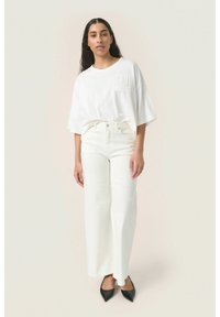 Hvid oversized t-shirt med lomme, parret med højtaljede hvide brede bukser og spidse sorte flade sko mod en neutral baggrund.