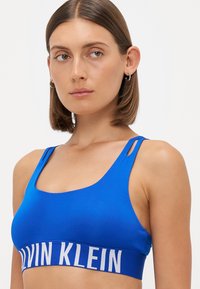 Koninklijk blauwe sportbh van rekbare stof, met een brede, getextureerde band met "CALVIN KLEIN" in vetgedrukte witte letters en smalle bandjes.