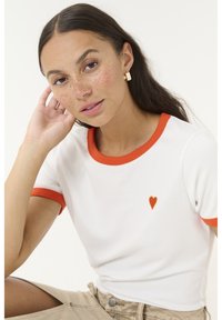 Weißes T-Shirt mit kurzen Ärmeln, orangefarbener Einfassung und einem kleinen roten Herzensymbol auf der Brust. Glatte Textur, taillierter Schnitt.