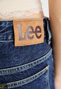 Blauwe denim jeans met een bruine leren patch waarop het 'Lee' logo is gestikt aan de achterste tailleband. De stof heeft een subtiele textuur en detaillering.