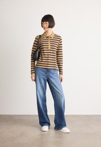 Donna che indossa una maglietta a maniche lunghe marrone e nera a righe, jeans larghi blu, sneakers bianche e tiene una borsa a tracolla nera, in piedi su un pavimento grigio.