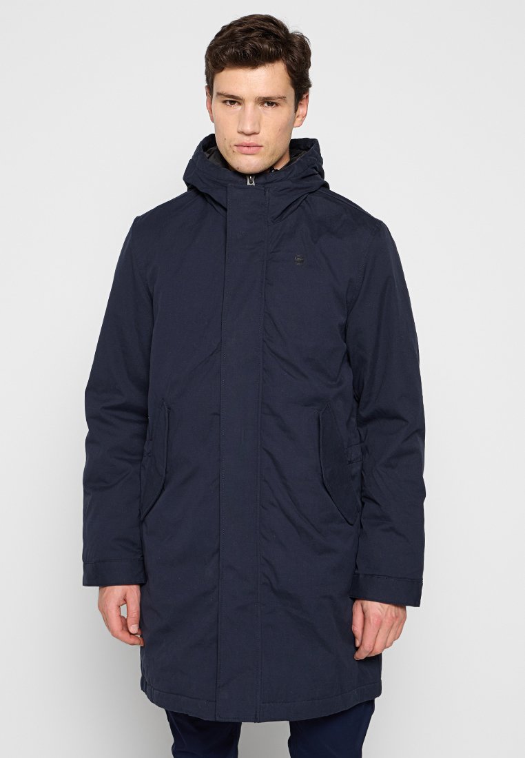 Parka bleu marine avec une fermeture éclair à l'avant, deux poches latérales, une capuche et une texture de tissu douce. Présente un logo discret sur la poitrine.