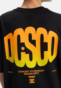 Zwarte katoenen t-shirt met grote gele en oranje gradient "DC SCO" tekst op de achterkant, met daaronder extra witte en oranje tekst.