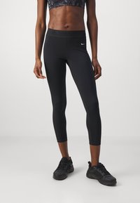 Kvinna står klädd i svarta korta leggings, svart sport-bh och svarta träningsskor framför en ljus, enfärgad bakgrund.