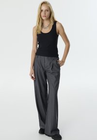 Zwarte nauwsluitende tanktop en grijze wijde broek. De broek heeft een geplooide voorkant en zijzakken; het model draagt zwarte schoenen en een kettinghalsband.