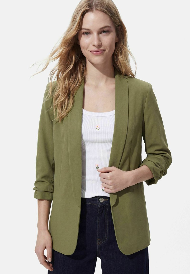 Femme aux longs cheveux blonds porte un blazer vert olive sur un débardeur blanc avec un petit motif floral et un jean foncé, souriante légèrement.