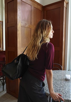 Schwarze Lederhandtasche, kastanienbraunes Strickoberteil und gestreifte Hose. Die Haare sind wellig, und die Umgebung besteht aus Holzverkleidung und einem Granittisch.