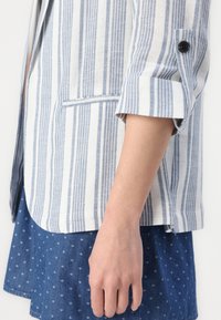 Blazer en lin rayé blanc et bleu, doté d'un poignet à un bouton, d'une poche ouverte et d'une coupe décontractée, associé à une jupe bleue à motifs.