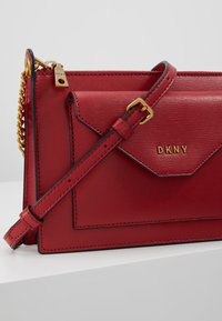 Czerwona skórzana torebka crossbody o teksturowanym wykończeniu, ze złotymi elementami, regulowanym paskiem oraz przednią kieszenią z klapą ozdobioną logo DKNY.