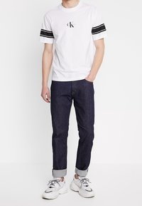 Valkoinen t-paita, jossa on mustat raidat hihoissa ja logo keskellä, yhdistettynä tummansinisiin farkkuihin ja valkoisiin tennareihin, joissa on harmaat yksityiskohdat.