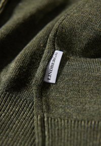Grön stickad tröja med texturerad tyg, prydd med en etikett som läser "LINDBERGH". Visar en ribbad fåll och krage.