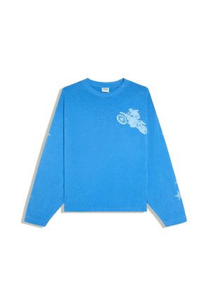 Giovane uomo con capelli scuri che indossa una camicia blu a maniche lunghe e pantaloncini abbinati con motivi a stelle, in piedi all’aperto vicino a una balaustra di pietra in un giardino.