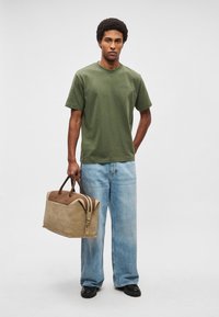 T-shirt de algodão verde, jeans azul-claro de ganga de corte largo, a segurar uma mala de couro bicolor bege e castanha com duas pegas e fecho éclair.