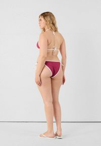 Bikini bordeaux avec des accents blancs ; design à col halter. Se noue autour du cou et sur les côtés. Le mannequin porte des sandales plates assorties. Texture lisse.