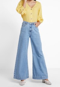 Lichtgele blouse met knopen en lange mouwen, gecombineerd met hooggevoerde, wijde lichtblauwe jeans en witte geperforeerde schoenen.