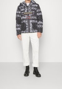 Man in een zwart-wit geblokte trui met Noorse vlag, witte opgerolde broek en zwarte veterschoenen.