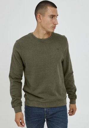 Felpa verde oliva con collo rotondo, polsini a costine e un logo discreto sul petto. Tessuto morbido e vestibilità rilassata. Jeans visibili.