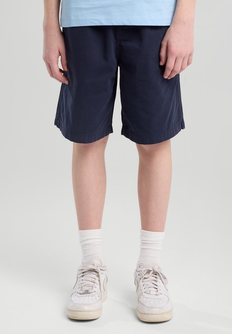 Pantalones cortos de color navy hechos de algodón, con un corte relajado y puños rematados, combinados con zapatillas blancas y calcetines blancos de canalé.