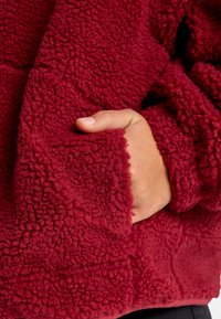 Giacca in pile bordeaux con materiale testurizzato e soffice. Presenta una mano in una tasca laterale che mostra un rivestimento interno morbido e accogliente.