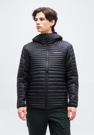 Giovane uomo che indossa una giacca imbottita nera con cappuccio e cerniera frontale e pantaloni cargo verde scuro, guarda verso sinistra.