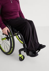 Μαύρο αναπηρικό καροτσάκι με πράσινες λεπτομέρειες. Άτομο με μαρoon μακρυμάνικη μπλούζα και μαύρο παντελόνι, φορώντας λαμπερά μαύρα παπούτσια, καθισμένο.