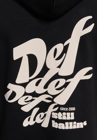 Svart hettegenser med stor hvit tekst i ulike stiliserte skrifttyper. Teksten inkluderer "Def," "siden 2006," og "fortsatt ballin.'" Mykt stoff.