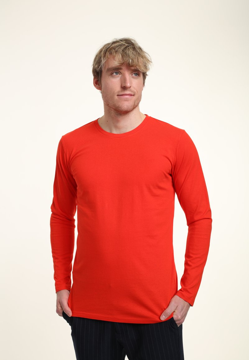 Röd långärmad T-shirt i mjukt tyg, med rund halsringning och figursydd design. Modellen poserar med händerna i fickorna.