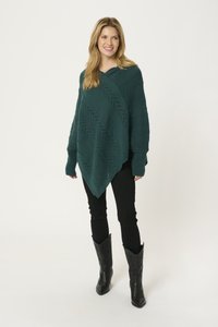 Poncho lavorato a maglia in tono verde acqua con scollo a V, caratterizzato da strisce diagonali testurizzate e dettagli a trama aperta, abbinato a pantaloni aderenti neri e stivali da cowboy neri.