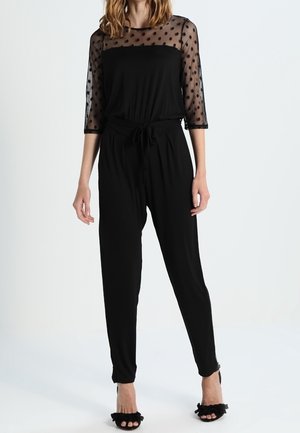 Vrouw draagt een zwarte jumpsuit met doorzichtige polkadotmouwen en zwarte open pumps met ruches, staand tegen een effen achtergrond.