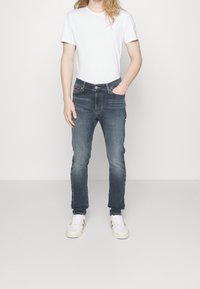 Úzké tmavě modré džíny s jemným vyblednutím, pěti kapsami a kontrastním prošitím. Kombinovány s jednoduchým bílým tričkem a bílými teniskami.